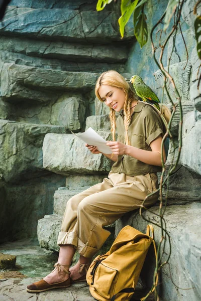 Heureuse jeune femme en costume safari avec perroquet à l'épaule et carte assise sur des rochers — Photo de stock