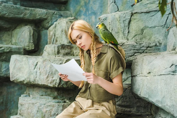 Belle jeune femme en costume safari avec perroquet à l'épaule et carte assise sur des rochers — Photo de stock