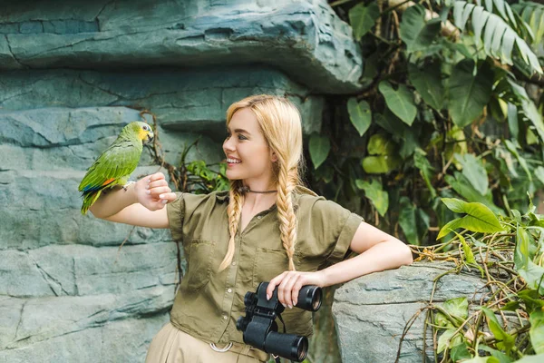 Attractive young woman in safari suit with parrot perching on arm in jungle — Stock Photo