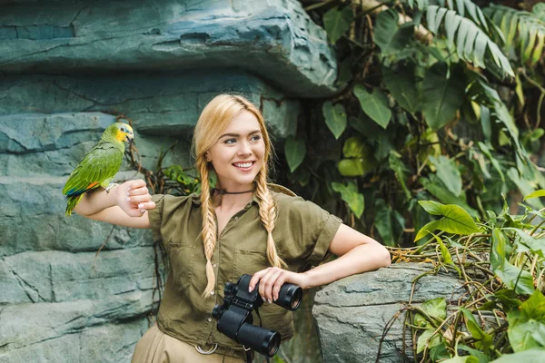 Happy young woman in safari suit with parrot perching on arm in jungle — Stock Photo