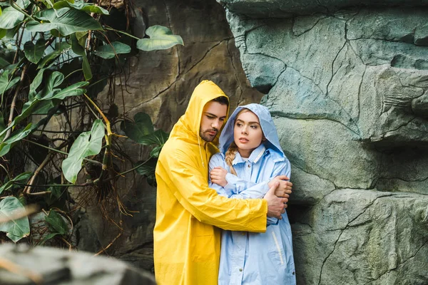Frozen young couple in raincoats embracing and trying to warm up under rocks in jungle — Stock Photo