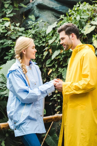 Beautiful young couple in raincoats holding hands at rainforest — Stock Photo