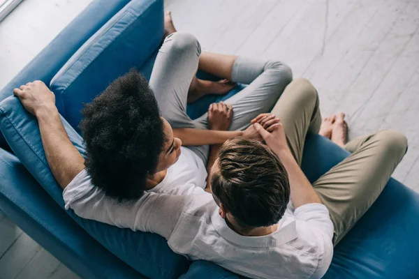 Pareja multiracial cogida de la mano mientras está sentada en el sofá - foto de stock