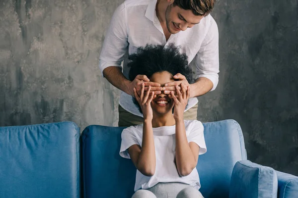 Mann verdeckt Augen einer afrikanisch-amerikanischen Freundin auf Sofa sitzend — Stockfoto
