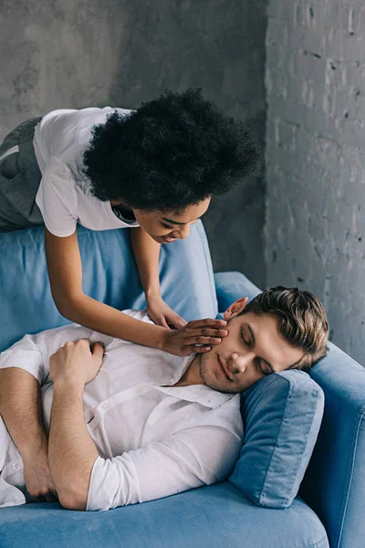 Mujer afroamericana tiernamente tocando la cara del hombre durmiendo en el sofá - foto de stock