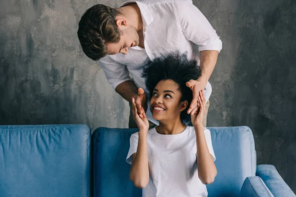 Hombre sorprendiendo a su novia afroamericana sentada en un sofá - foto de stock