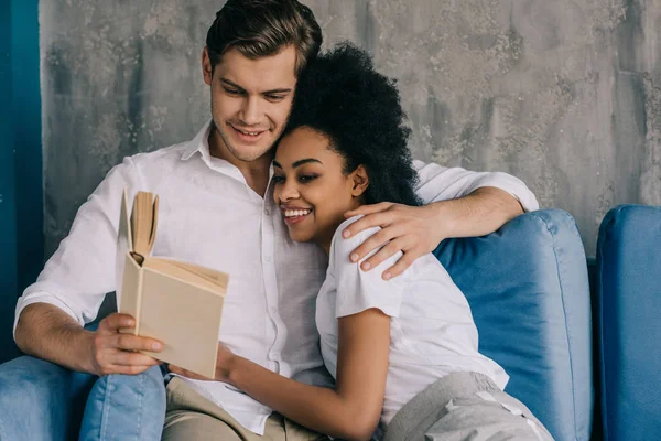 Vielrassig umarmendes junges Paar liest Buch auf Sofa — Stockfoto