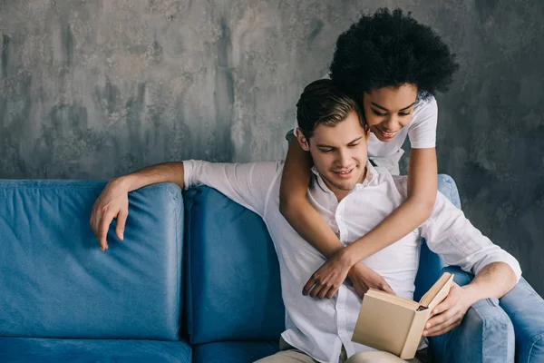 Novio y novia multirraciales discutiendo libro juntos en sofá - foto de stock