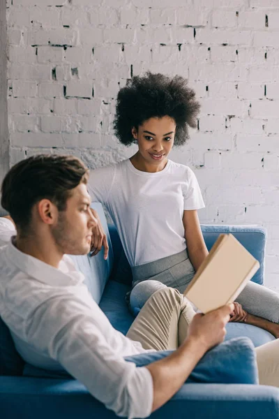 Multiracial boyfriend and girlfriend reading book on sofa at home — Stock Photo