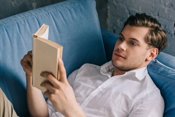 Jeune homme relaxant à la maison sur canapé avec livre — Photo de stock