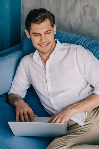Smiling man working on laptop while lying on sofa — Stock Photo