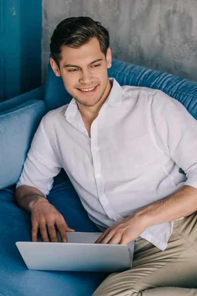 Homme souriant travaillant sur un ordinateur portable tout en étant couché sur un canapé — Photo de stock