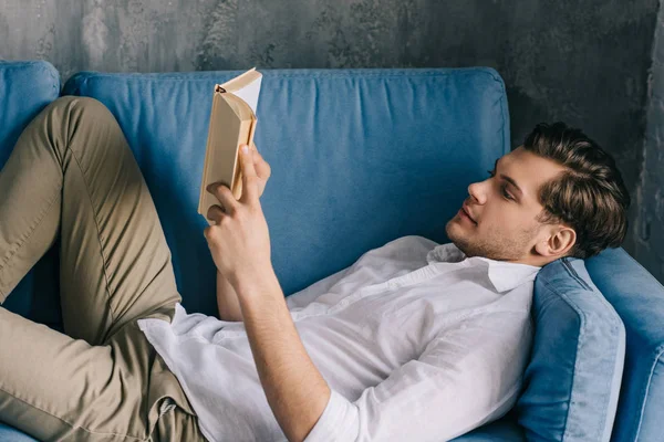 Junger Mann liest Buch, während er auf dem Sofa liegt — Stockfoto