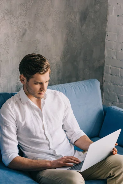 Homme tapant sur le clavier de l'ordinateur portable sur ses genoux tout en étant assis sur le canapé — Photo de stock