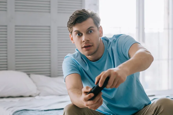 Überraschter junger Mann mit Joystick spielt Videospiel, während er auf dem Bett sitzt — Stockfoto