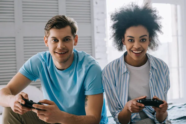 Multiracial couple with joysticks playing video game while sitting on bed — Stock Photo
