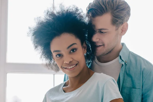 Joven abrazando a novia afroamericana por ventana en la habitación - foto de stock