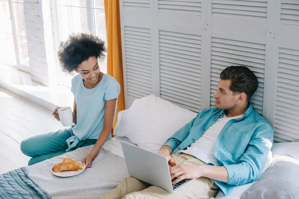 African american woman having breakfast by her boyfriend in bed with laptop — Stock Photo