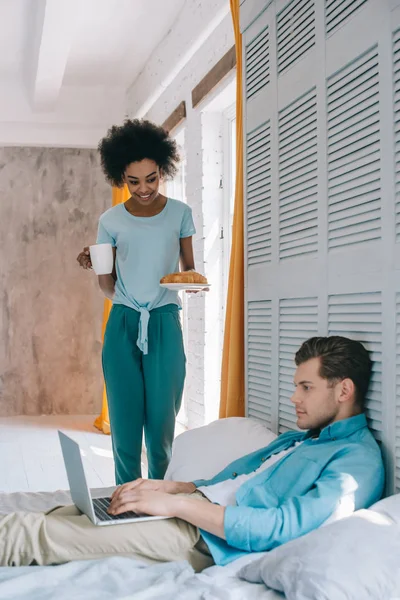 African american girl standing with breakfast by her boyfriend in bed with laptop — Stock Photo