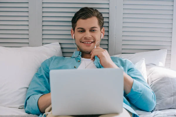 Hombre joven en auriculares usando el ordenador portátil mientras está acostado en la cama - foto de stock