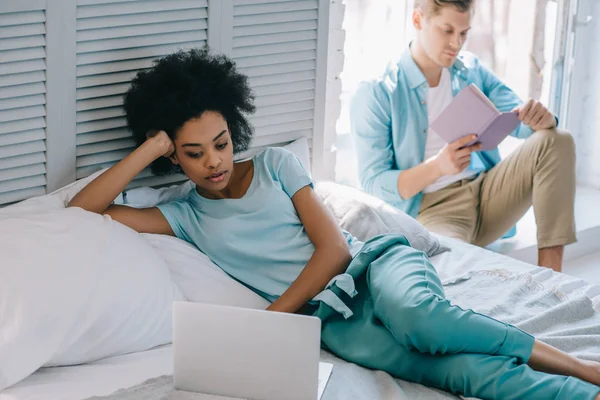 Afrikanerin benutzt Laptop, während Mann zu Hause Buch liest — Stockfoto