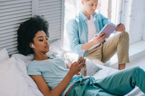 Relaxante mulher ouvir música no telefone enquanto o homem ler livro no peitoril da janela — Fotografia de Stock