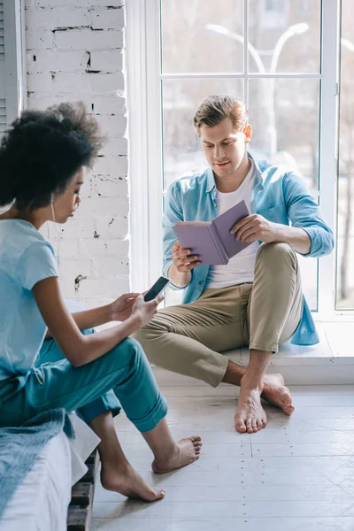 Жінка слухати музику на телефон під час людина читання книги на підвіконня — стокове фото