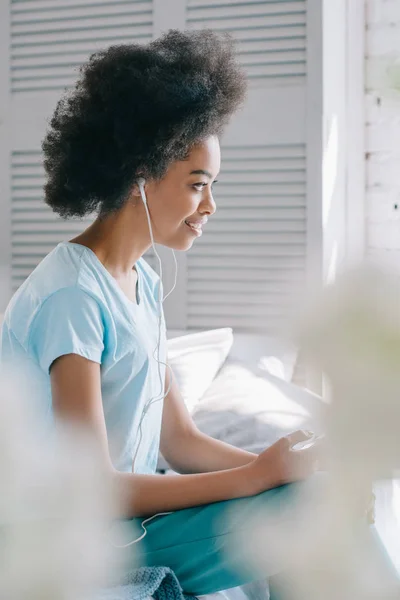 Menina americana africana feliz sentado na cama e ouvir música no telefone — Fotografia de Stock