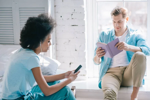 Afrikanerin hört Musik am Telefon, während Mann zu Hause Buch liest — Stockfoto