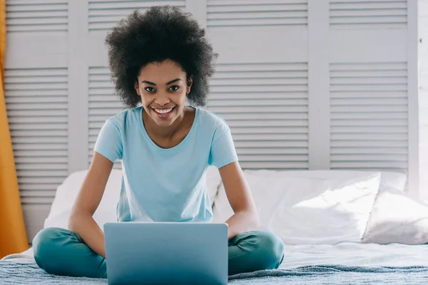 Sonriendo chica afroamericana utilizando el ordenador portátil mientras está sentado en la cama - foto de stock