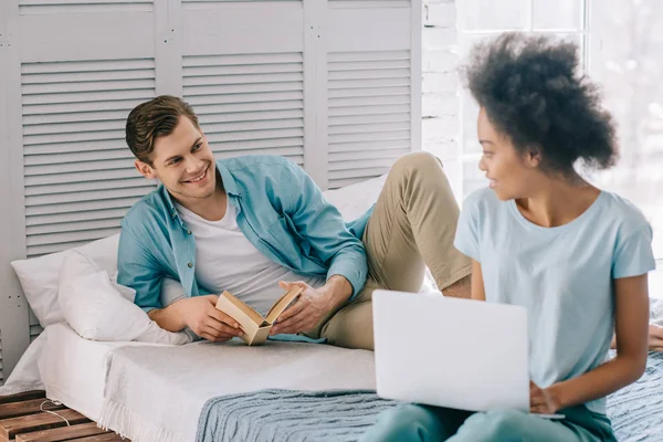 Africano americano chica sosteniendo portátil mirando hombre con lectura en cama en casa - foto de stock