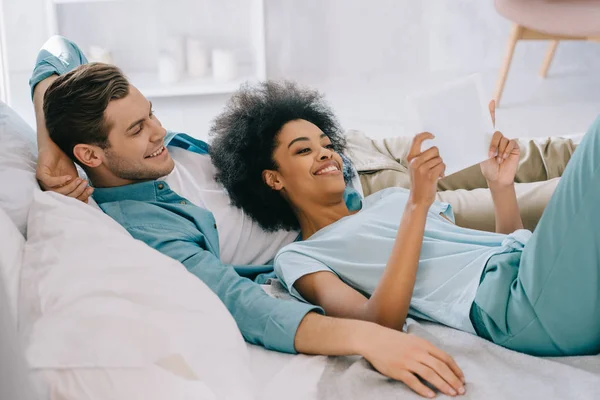 Multiracial couple looking at tablet screen while lying in bed — Stock Photo