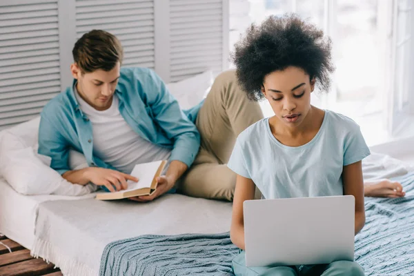 Africano menina americana usando laptop enquanto o homem lendo livro na cama em casa — Fotografia de Stock