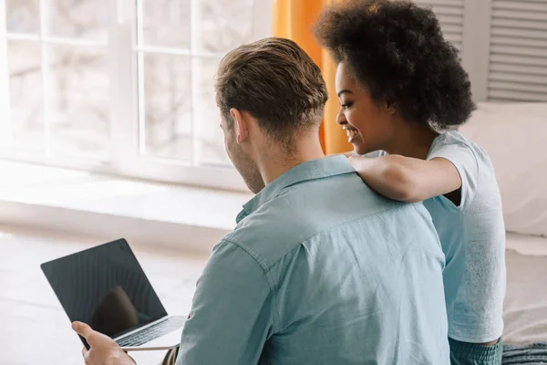 Pareja multiracial mirando a la pantalla del ordenador portátil mientras está sentado en la cama — Stock Photo