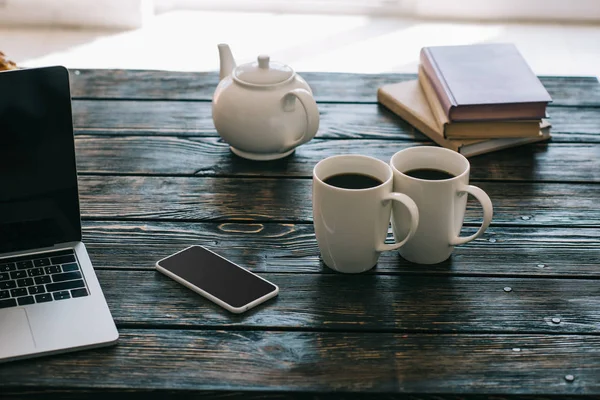 Tasses et théière sur table en bois avec ordinateur portable et smartphone — Photo de stock