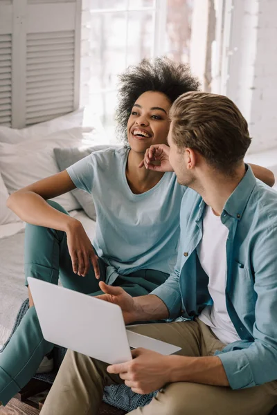 Multiracial boyfriend and girlfriend using laptop while sitting on bed — Stock Photo