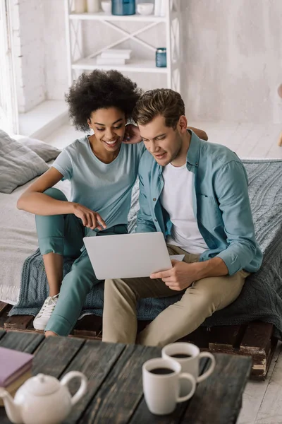 Smiling multiracial couple looking at laptop screen while sitting on bed — Stock Photo