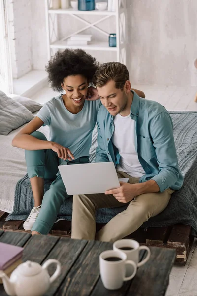 Lächelndes multiethnisches Paar, das auf Laptop-Bildschirm schaut, während es im Bett sitzt — Stockfoto