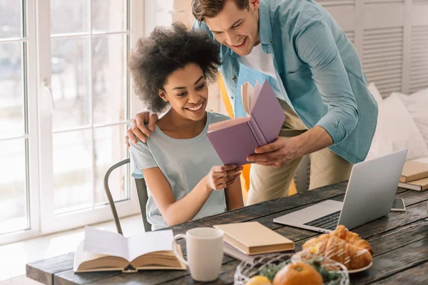 Vielrassiges junges Paar liest Buch an Tisch mit Laptop — Stockfoto