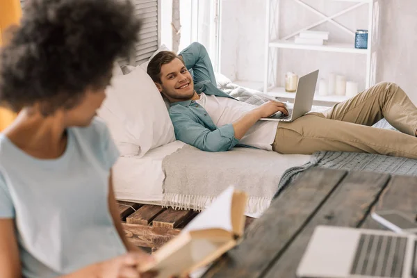 Африканская американка читает книгу за мужчиной, лежащим на кровати с ноутбуком — стоковое фото