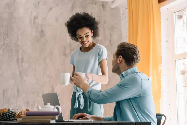 Pareja multirracial tomando café de la mañana por la mesa con el ordenador portátil - foto de stock