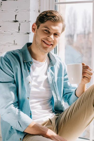 Hombre joven relajado sosteniendo la taza de café mientras está sentado en el alféizar de la ventana - foto de stock
