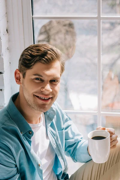 Hombre sonriente sosteniendo una taza de café mientras está sentado en el alféizar de la ventana - foto de stock