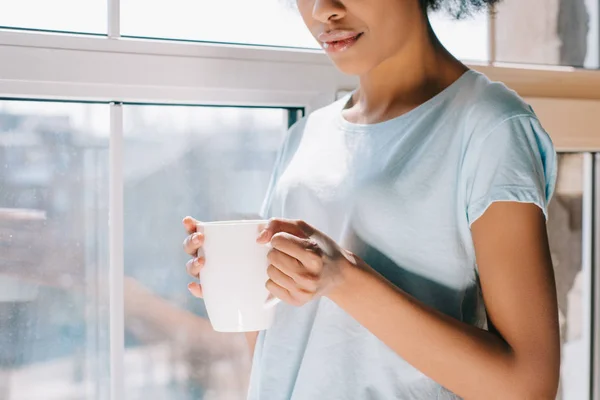 Close-up vista de xícara de café em mãos de menina americana africana em pé pela janela — Fotografia de Stock