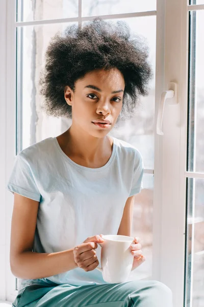 African american woman in lounge wear holding cup of coffee while sitting on windowsill — Stock Photo