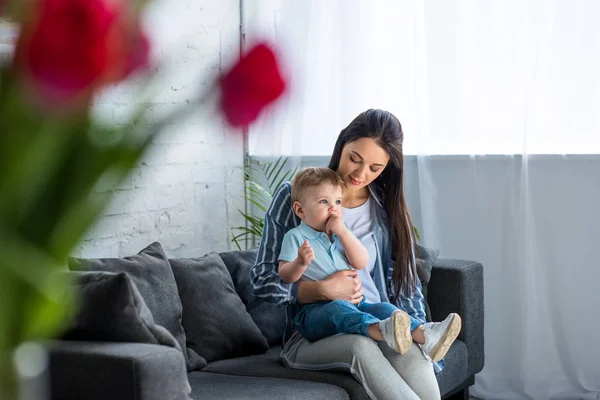 Foco seletivo da mãe com menino adorável em mãos sentadas no sofá em casa — Fotografia de Stock