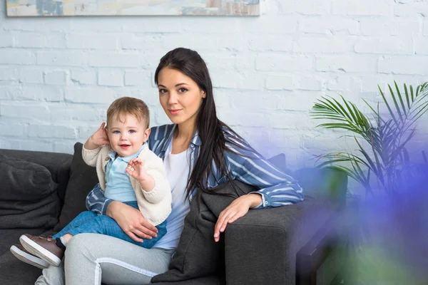 Mère avec adorable bébé garçon sur les mains assis sur le canapé à la maison — Photo de stock