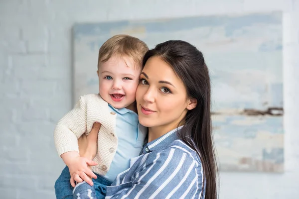 Porträt einer jungen Mutter, die zu Hause einen fröhlichen, entzückenden Jungen in den Händen hält — Stockfoto