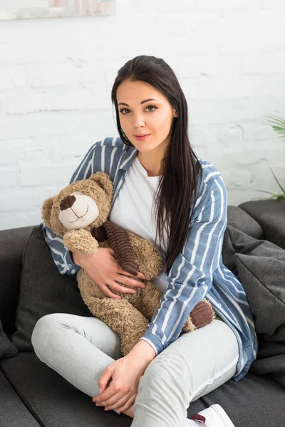 Portrait de femme souriante avec un ours en peluche reposant sur un canapé à la maison — Photo de stock