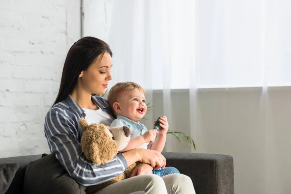 Jovem mãe e pequeno bebê sorridente com ursinho de pelúcia sentado no sofá em casa — Fotografia de Stock